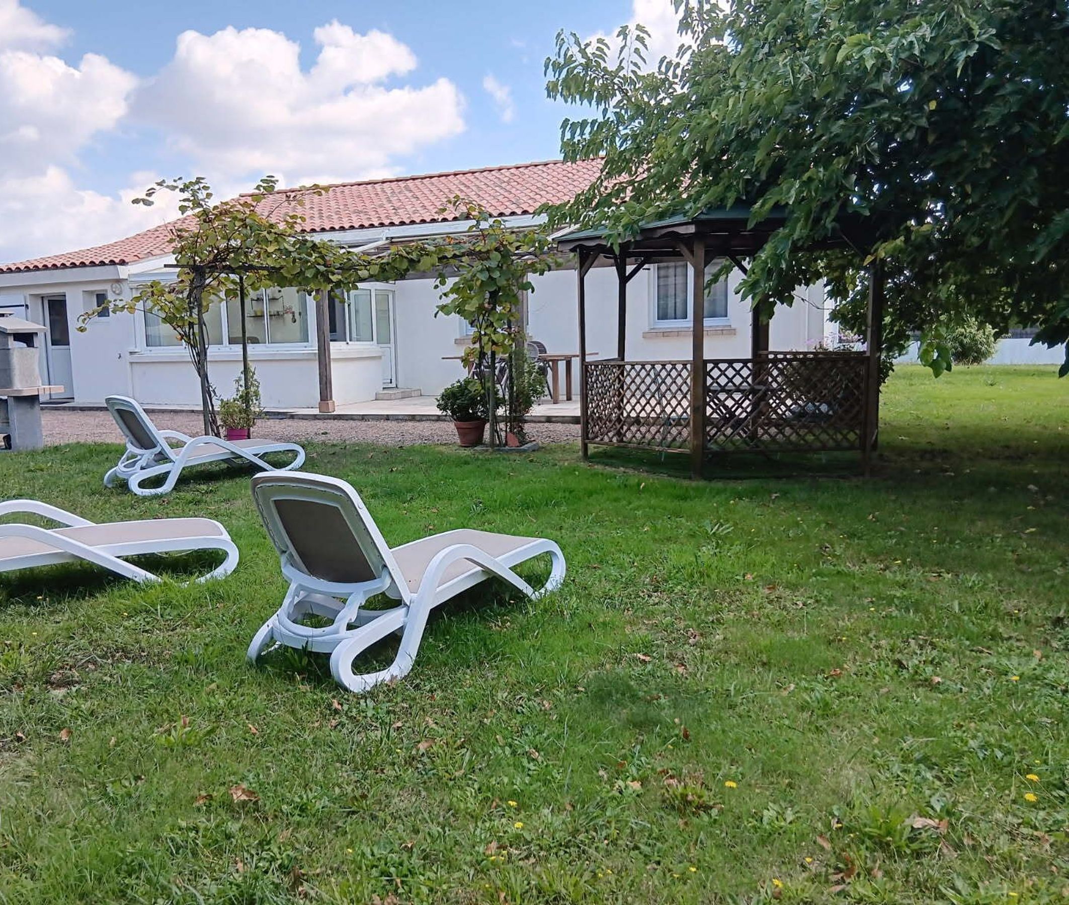 Un espacio cubierto de césped con tres sillas reclinables blancas, un edificio blanco con techo de tejas rojas y una pérgola de madera.