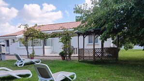 Une zone herbeuse avec trois chaises longues blanches, un bâtiment blanc doté d’un toit recouvert de tuiles rouges et une pergola en bois.
