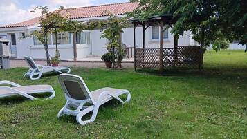 Un espacio cubierto de césped con tres sillas reclinables blancas, un edificio blanco con techo de tejas rojas y una pérgola de madera.