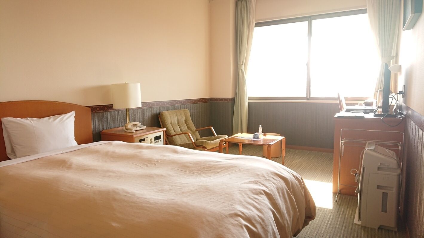A hotel room with a large bed, a chair, a desk with a lamp, and a window with curtains.