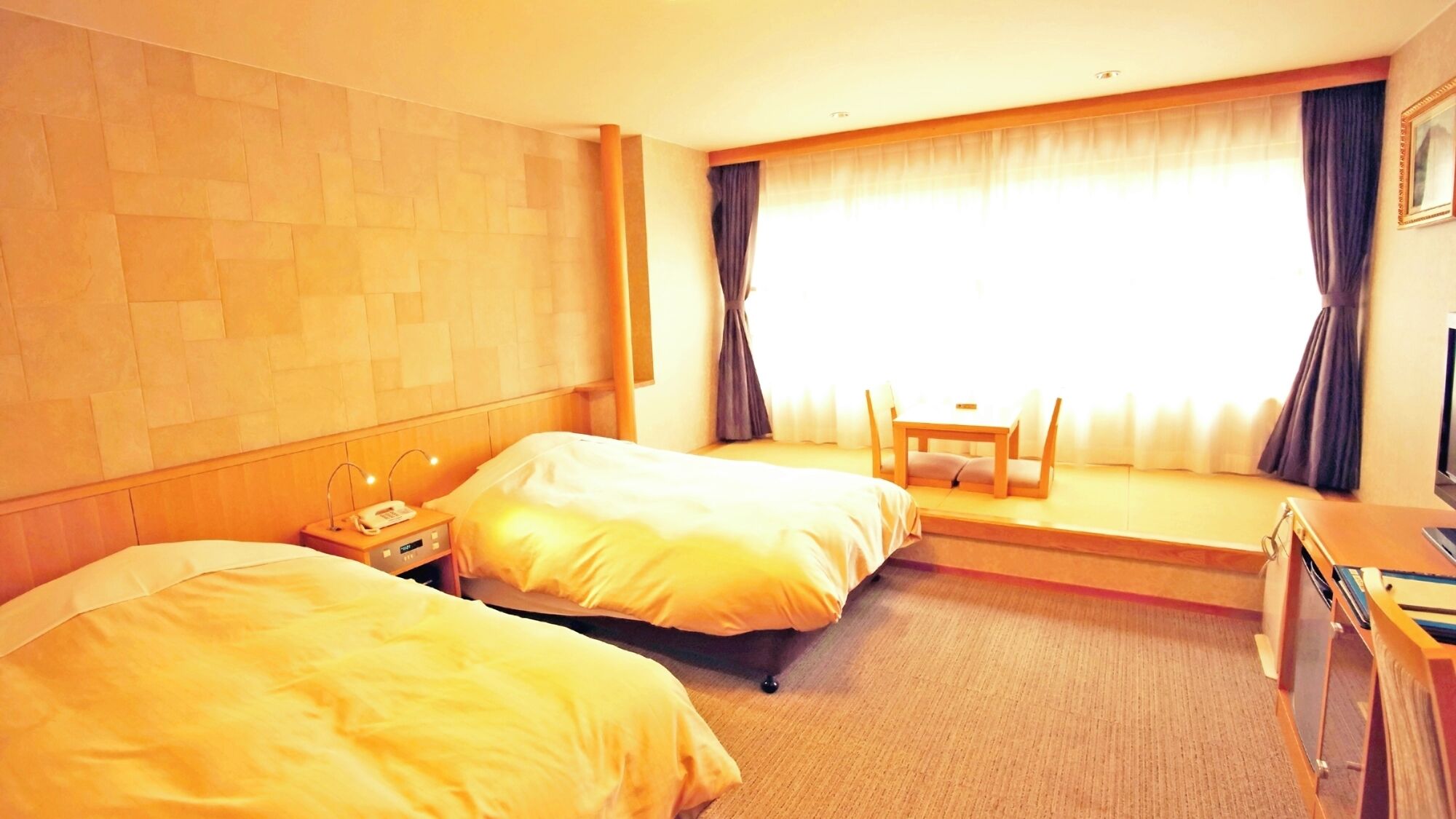 Habitación de hotel con dos camas, una mesita pequeña, una silla, un escritorio y una ventana con cortinas.