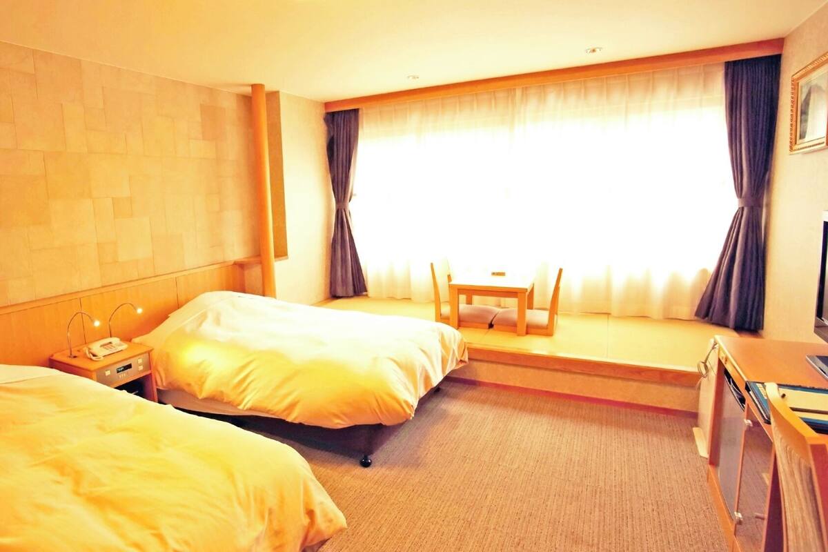 Habitación de hotel con dos camas, una mesita pequeña, una silla, un escritorio y una ventana con cortinas.