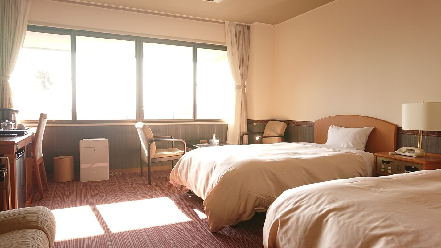 A hotel room with two beds, a desk, a chair, a lamp, and a window.