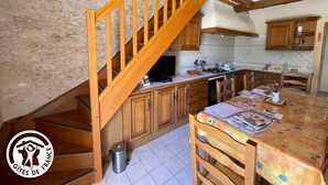 A kitchen with wooden cabinets, a tiled floor, a dining table with chairs, a wooden staircase, and a trash bin. - Charming Riverside House with Canoes, Garden, and Nearby Market in Marais Poitevin (Damvix)
