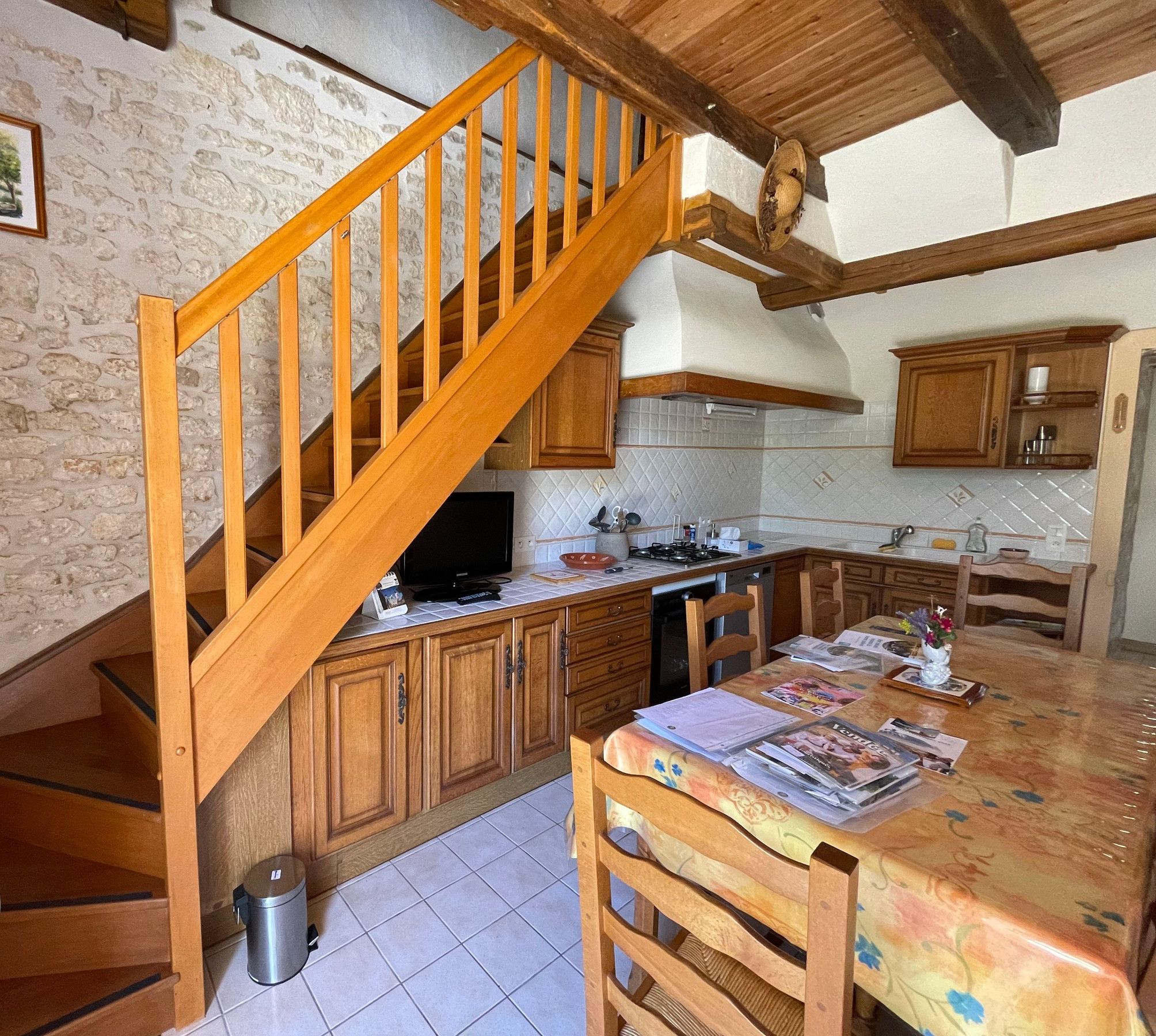 Une cuisine avec des meubles de rangement en bois, un sol carrelé, une table à manger avec des chaises, un escalier en bois et une poubelle.