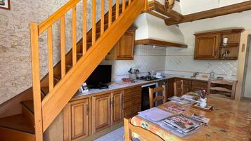 Une cuisine avec des meubles de rangement en bois, un sol carrelé, une table à manger avec des chaises, un escalier en bois et une poubelle.
