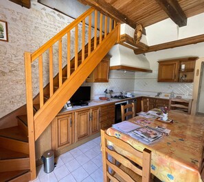 A kitchen with wooden cabinets, a tiled floor, a dining table with chairs, a wooden staircase, and a trash bin.