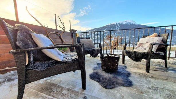 Un'accogliente area salotto all'aperto con sedie e cuscini in vimini, un braciere in metallo e vista su una montagna innevata.