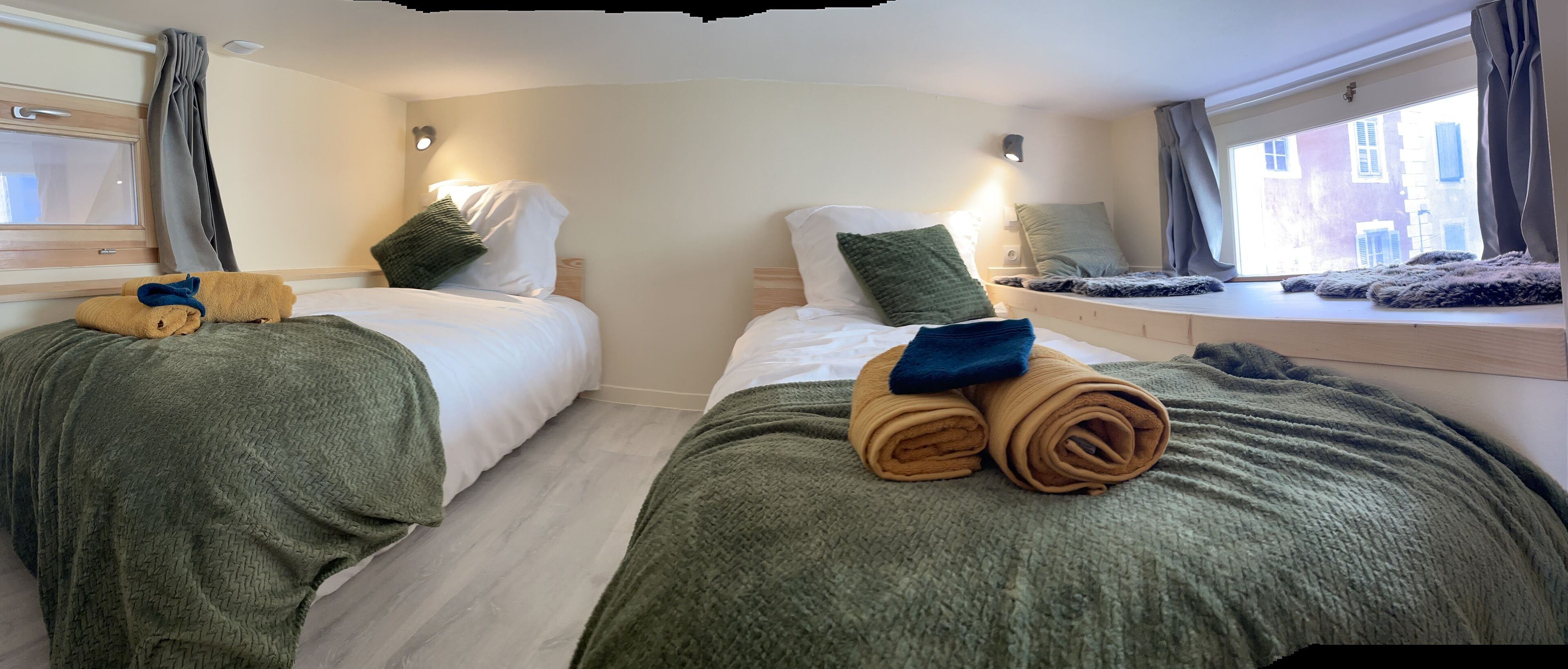 A hotel room with two beds, each with a green blanket and a rolled towel. There are pillows on the beds, and a window with curtains.