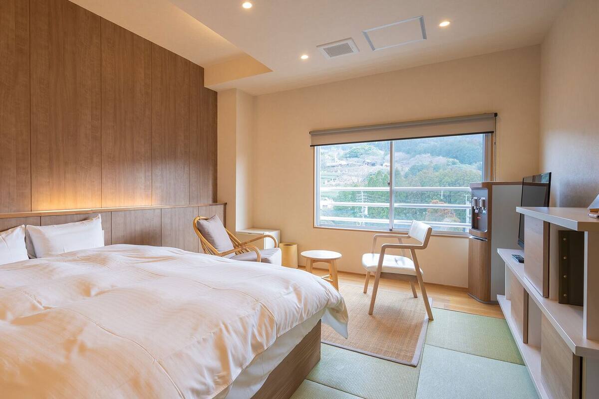 A hotel room with a large bed, a wooden headboard, a small table, a chair, a television, and a window with a view.