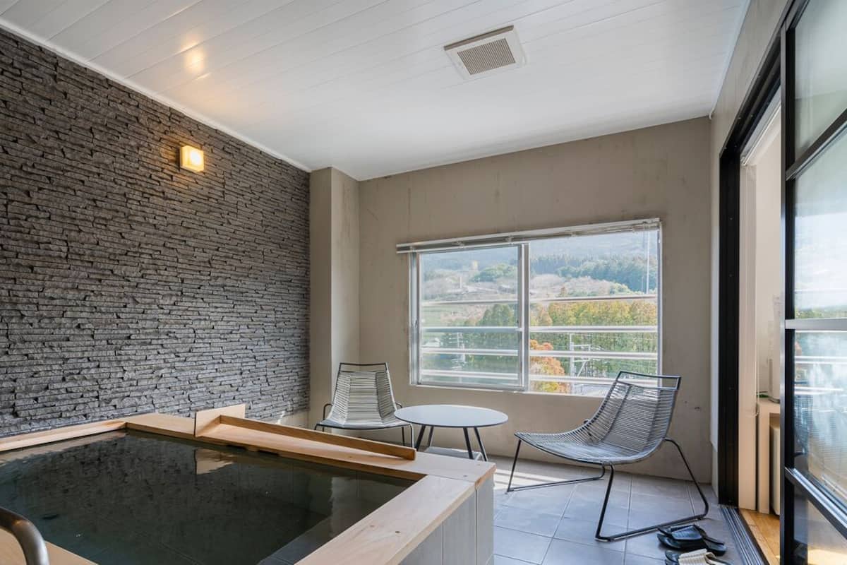 A modern room with a stone wall, a hot tub, a chair, and a window with a view.