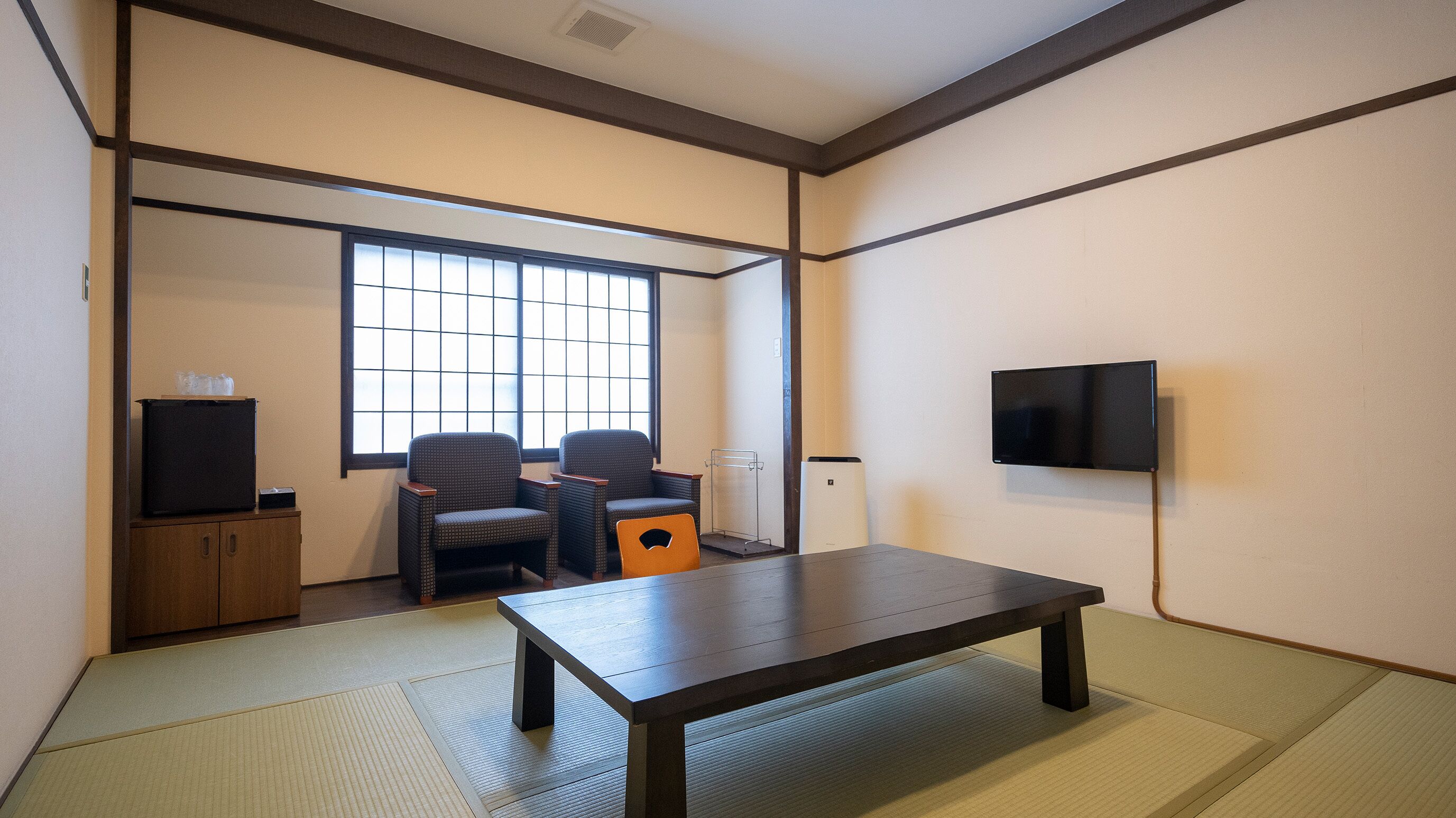 Ein Zimmer mit Tatami-Böden, einem niedrigen Tisch, zwei Stühlen, einem Fernseher und einem Schrank.