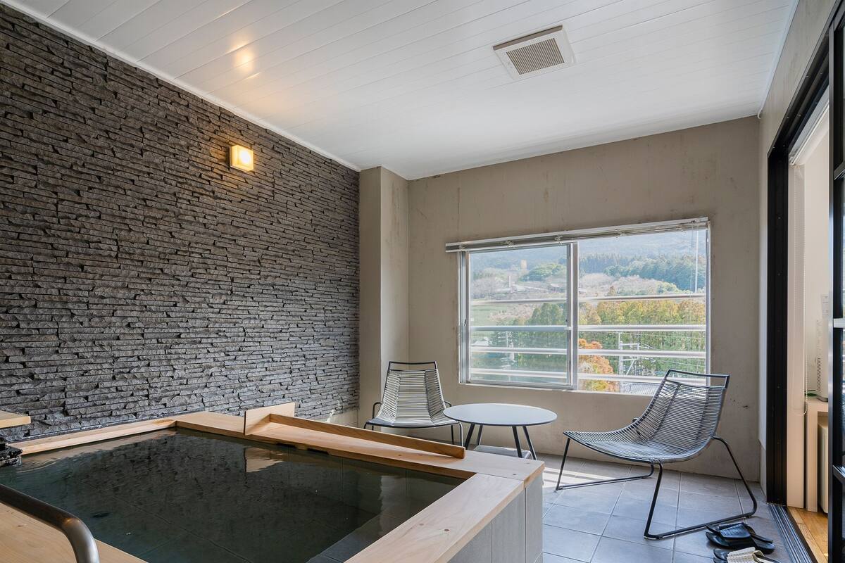 A spa area with a stone wall, a hot tub, a chair, and a view of the outdoors.