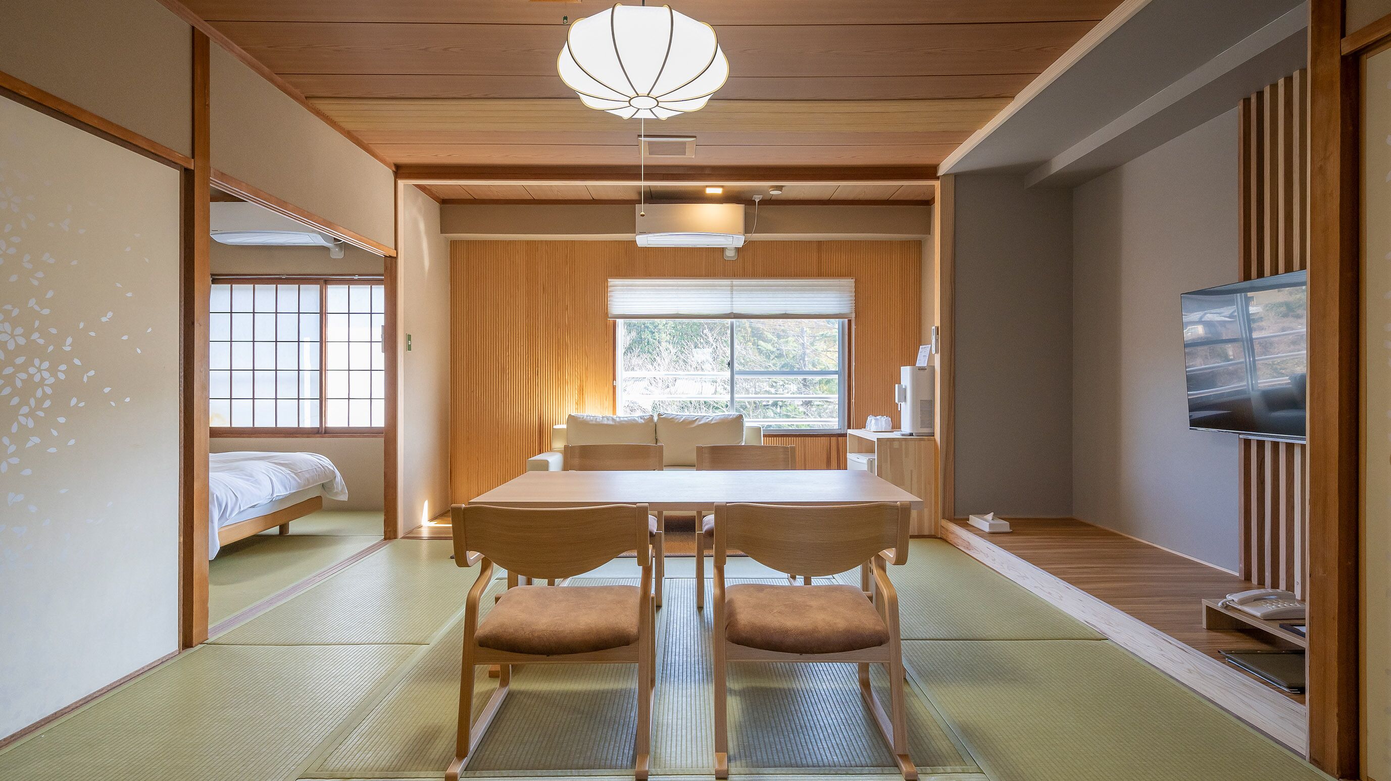 Ein traditionelles japanisches Zimmer mit Tatami-Böden, einem niedrigen Tisch mit Stühlen, einem Fernseher und einer Schiebetür zu einem Schlafbereich.