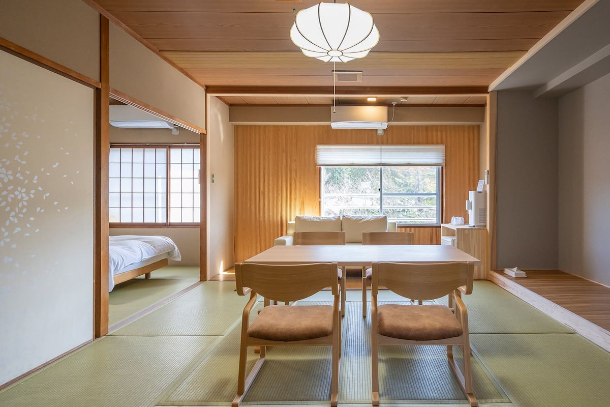 A traditional Japanese-style room with tatami flooring, a low table with chairs, a television, and a sliding door leading to a sleeping area.