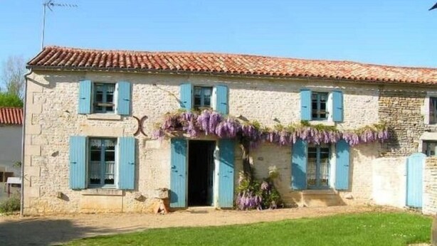 Ein Gebäude aus Stein mit einem Ziegeldach, blauen Fensterläden und einem kleinen Garten mit violetten Blumen.