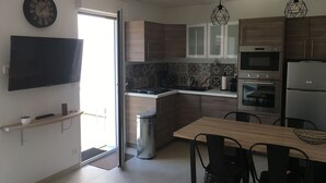 A modern kitchen with wooden cabinets, a built-in oven, a microwave, and a stainless steel refrigerator. There is a dining table with chairs, a trash can, and a clock on the wall.