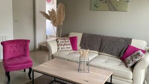 A living room with a white sofa, a pink armchair, and a coffee table. There is a painting on the wall and a door in the background.