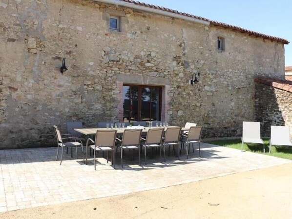 A stone building with a tiled roof, a large table with chairs, and a paved patio area. - 18th Century Lodge, Sleeps 10 (Les Herbiers)