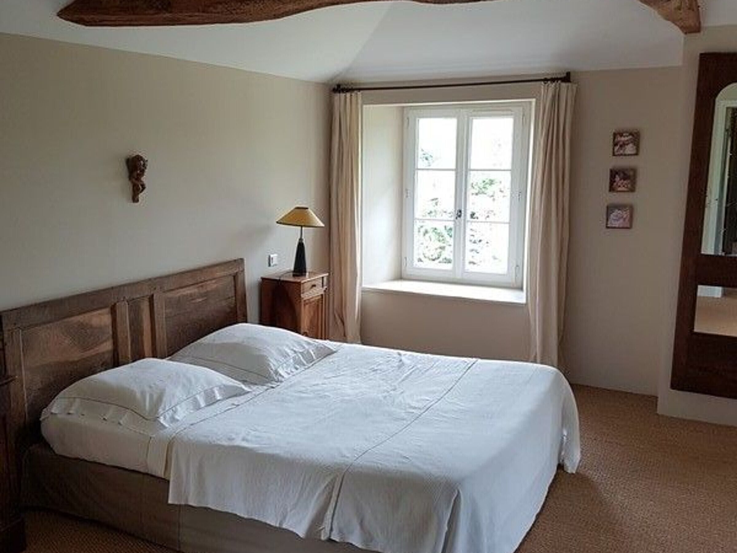 Una cama bien hecha con sábanas blancas en una habitación con cabecera de madera, una mesita de noche con lámpara y una ventana con cortinas.