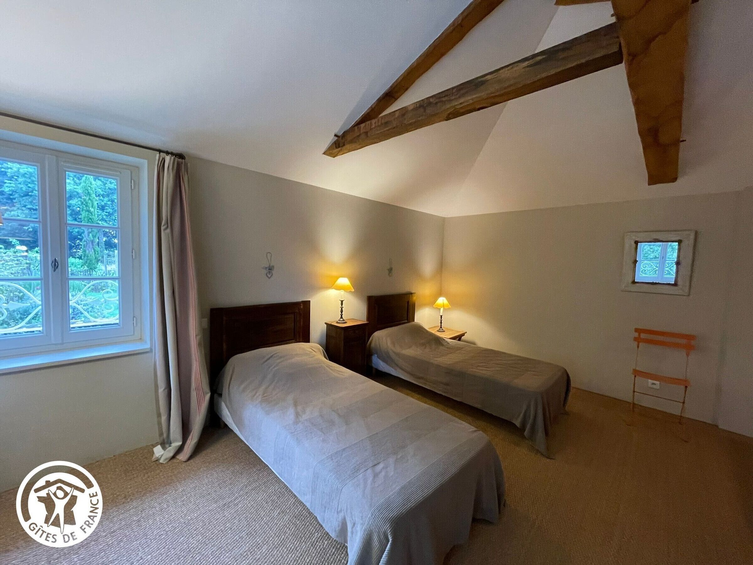 A bedroom with two beds, a window with curtains, a small table with a lamp, and a wooden beam on the ceiling.