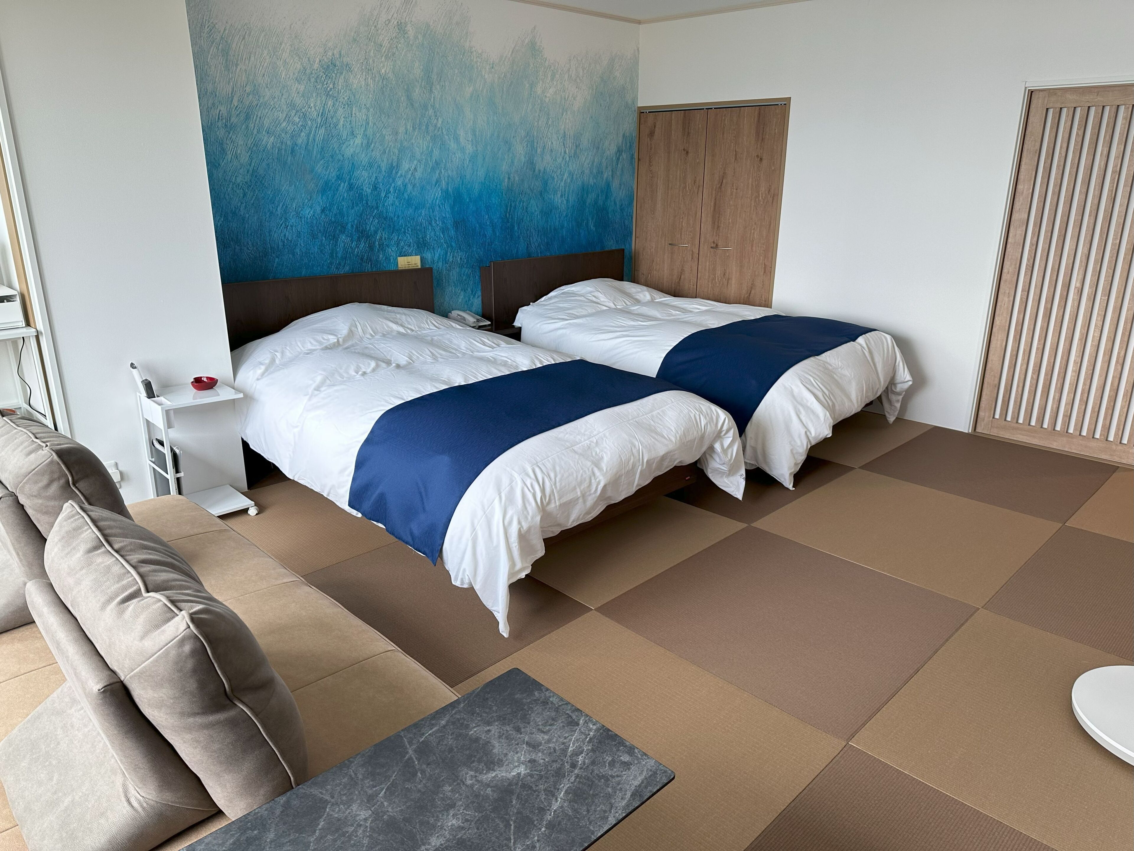A hotel room with two beds, a sofa, a small table, and a wall with a blue abstract design.