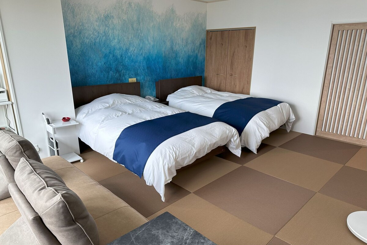 A hotel room with two beds, a sofa, a small table, and a wall with a blue abstract design.
