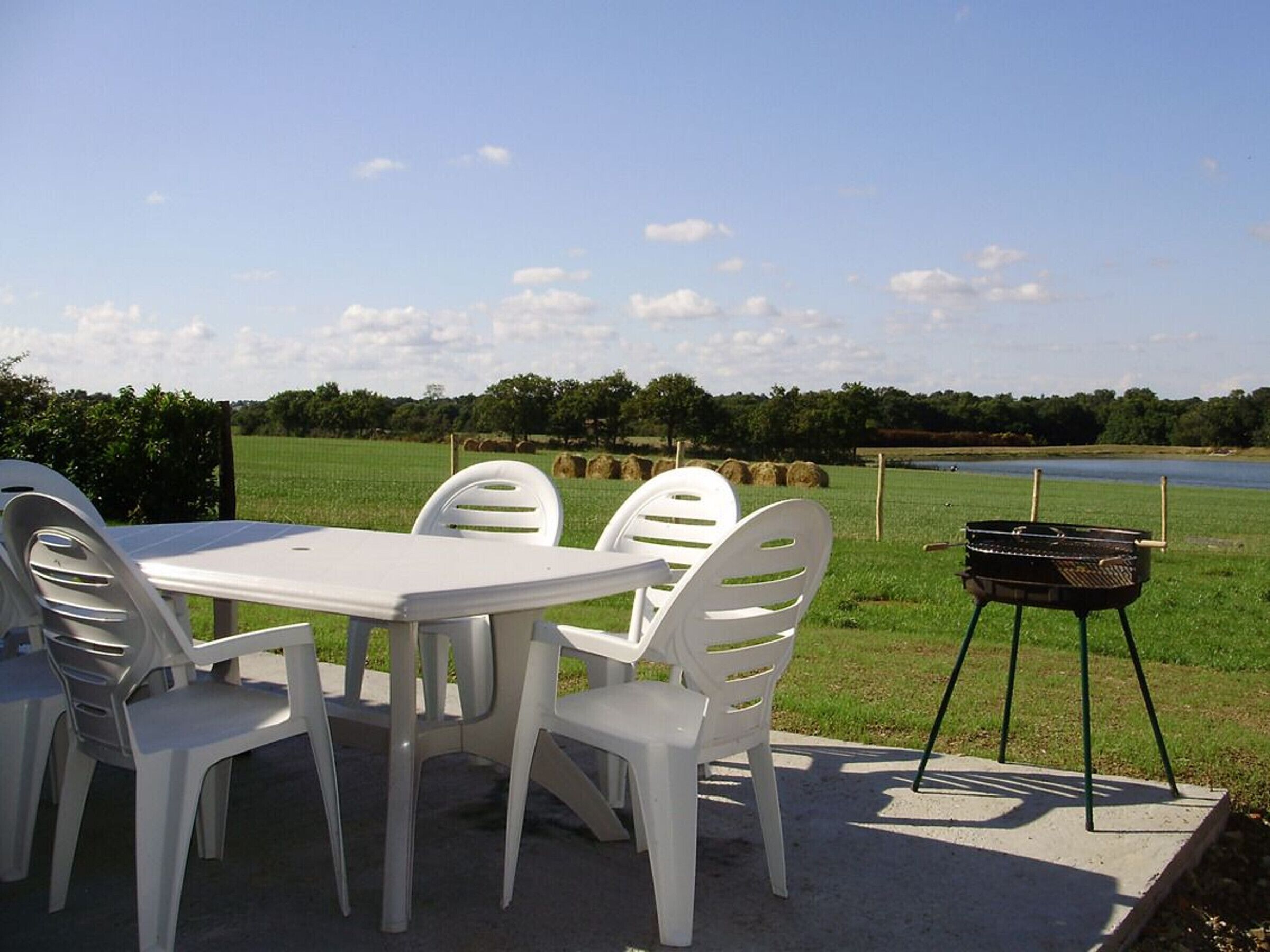 A picnic setup with a table and chairs, a grill, and a view of a lake and fields.