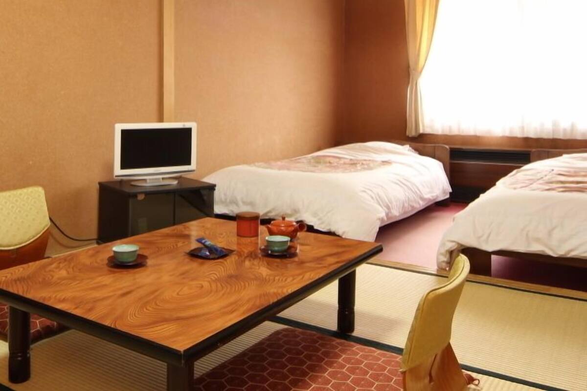 Quarto de hotel com duas camas, uma mesa de madeira, televisão e janela com cortinas.