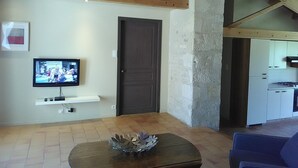 A living room with a television, a door, and a kitchen in the background. - Charming 18th-Century Cottage with Pool, Terrace & Garden in the French Countryside (Auchay-sur-Vendée)
