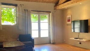 A spacious living room with a large window, a flat-screen TV mounted on the wall, and a wooden ceiling. - Charming 18th-Century Cottage with Pool, Terrace & Garden in the French Countryside (Auchay-sur-Vendée)