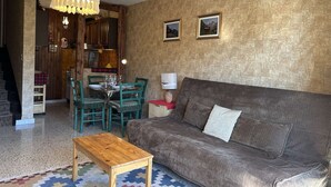 A cozy living room with a brown sofa, a wooden coffee table, and a patterned rug. There is a dining area with a table set for four, chairs, and a chandelier. The walls are textured, and there are two framed pictures on the wall. - Cozy Studio for 4 in Montgenèvre – Balcony, Ski Locker, Close to Slopes & Shops (Montgenèvre)