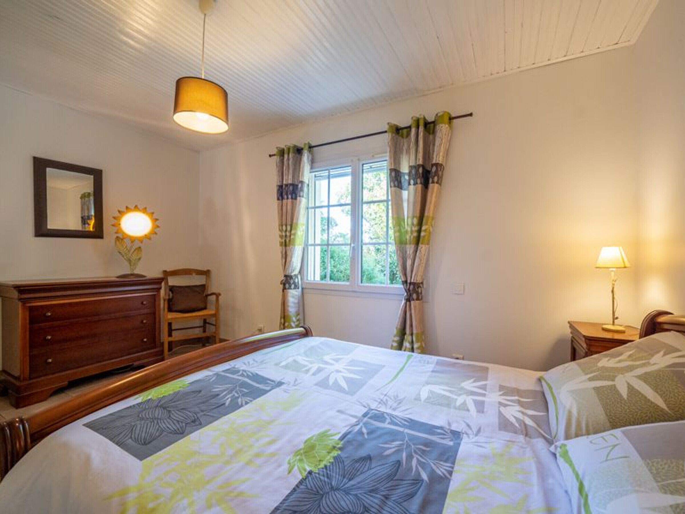A bedroom with a bed, a dresser, a chair, a window with curtains, and a lamp.