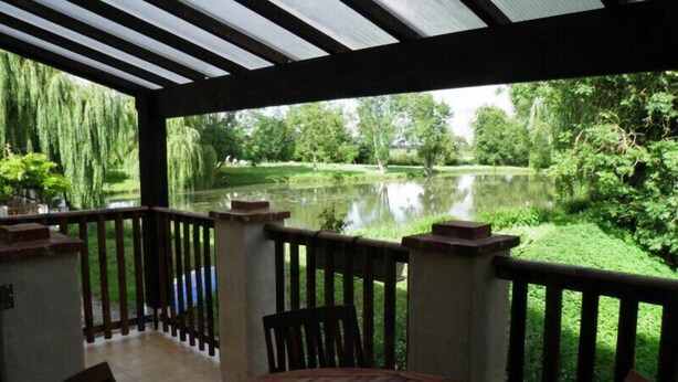 A covered outdoor area with a wooden table and chairs, overlooking a pond and lush greenery. - Charming Watermill Gîte: 6BR, Fireplace, Private Garden, Pond, Terrace, WiFi, Linens Included (Foussais-Payré)