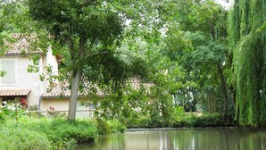 A serene riverside scene with a building featuring a tiled roof and a balcony. - Charming Watermill Gîte: 6BR, Fireplace, Private Garden, Pond, Terrace, WiFi, Linens Included (Foussais-Payré)