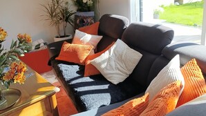 Een moderne woonkamer met een donkergrijze bank, oranje en witte kussens, een glazen salontafel en uitzicht op een groene tuin door grote ramen.