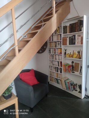 A modern living room with a staircase, a bookshelf filled with books, a grey armchair with a red pillow, and a small table with a plant.