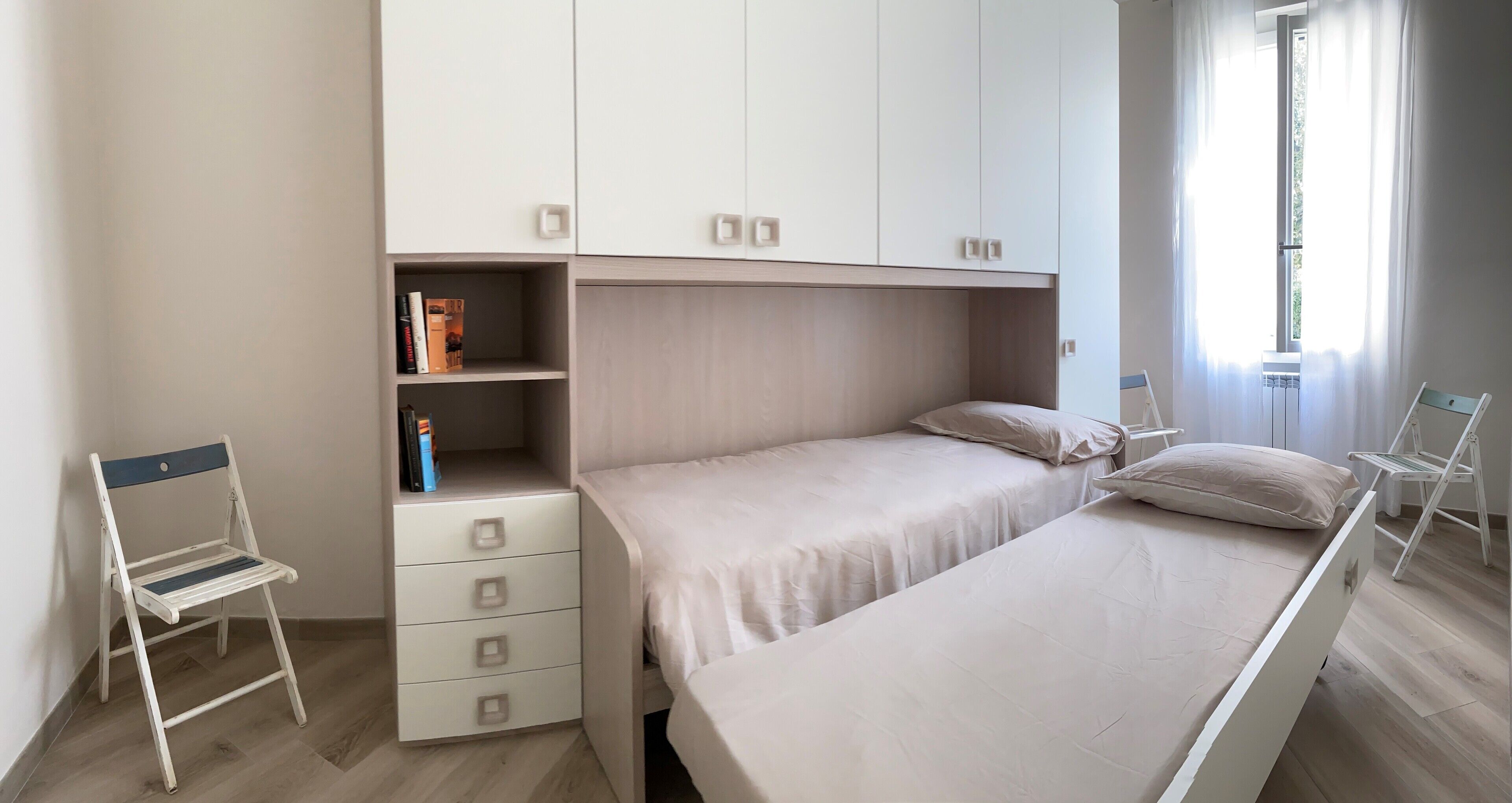 Ein Schlafzimmer mit einem Bett, einem Sessel, einem kleinen Bücherregal und einem Fenster mit Vorhängen.