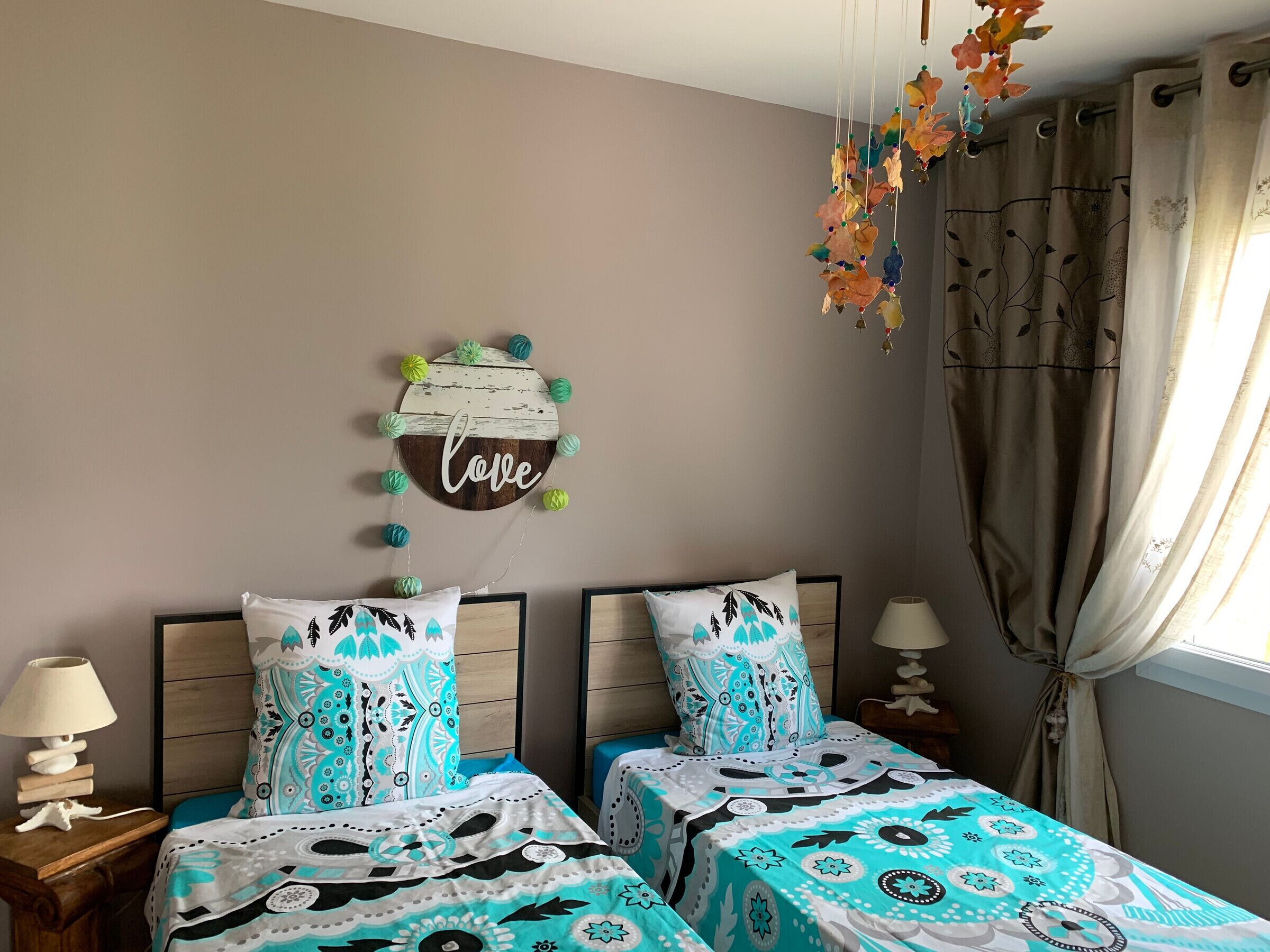 A bedroom with two beds, a wall decoration with the word "love" and colorful pom-poms, a lamp on the nightstand, and a window with curtains.