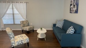 A living room with a blue sofa, patterned armchairs, a small white table, and a window with blinds.