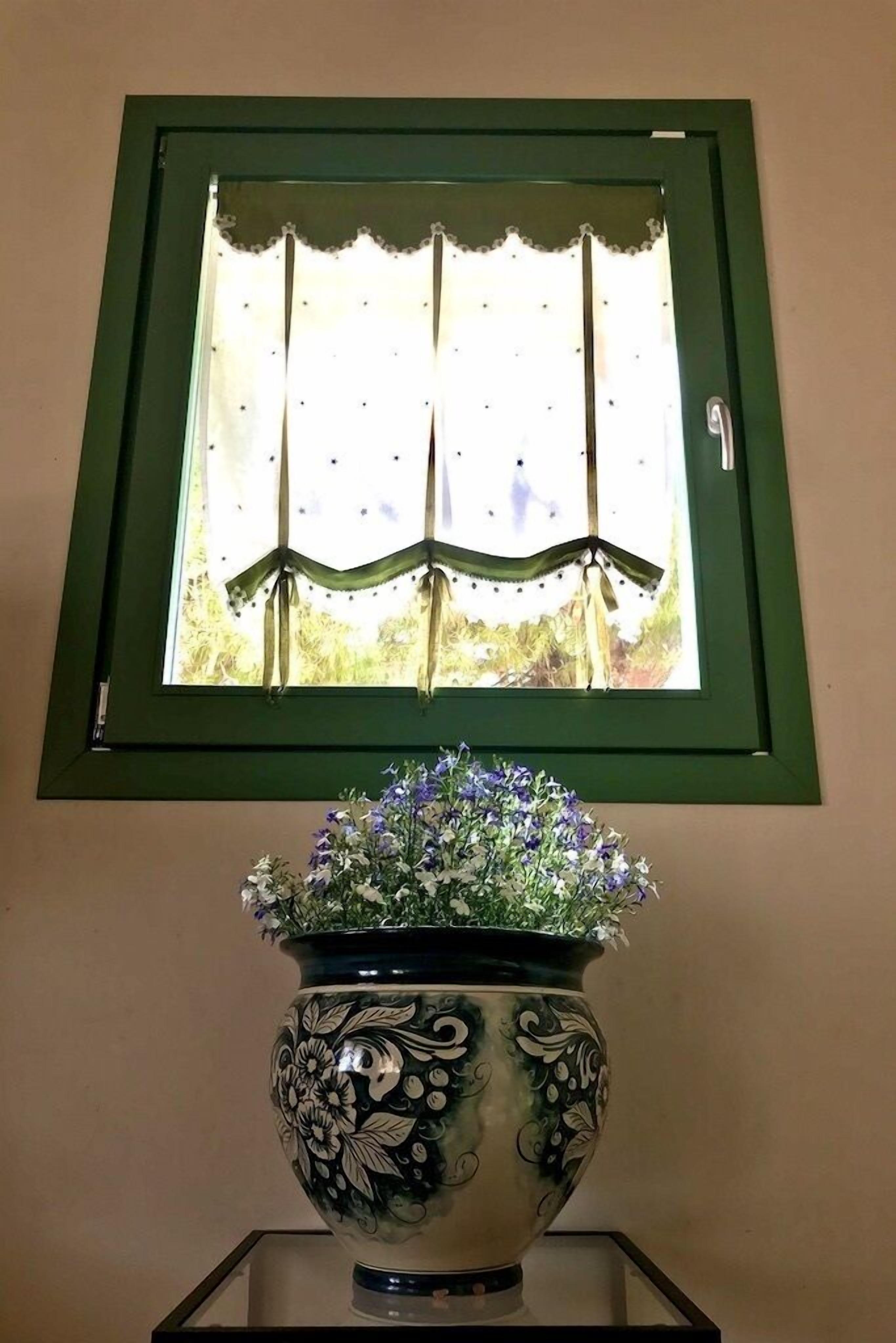 Una finestra con cornice verde e una vista decorativa su un paesaggio. Sotto la finestra, un vaso bianco con motivo floreale e fiori viola.
