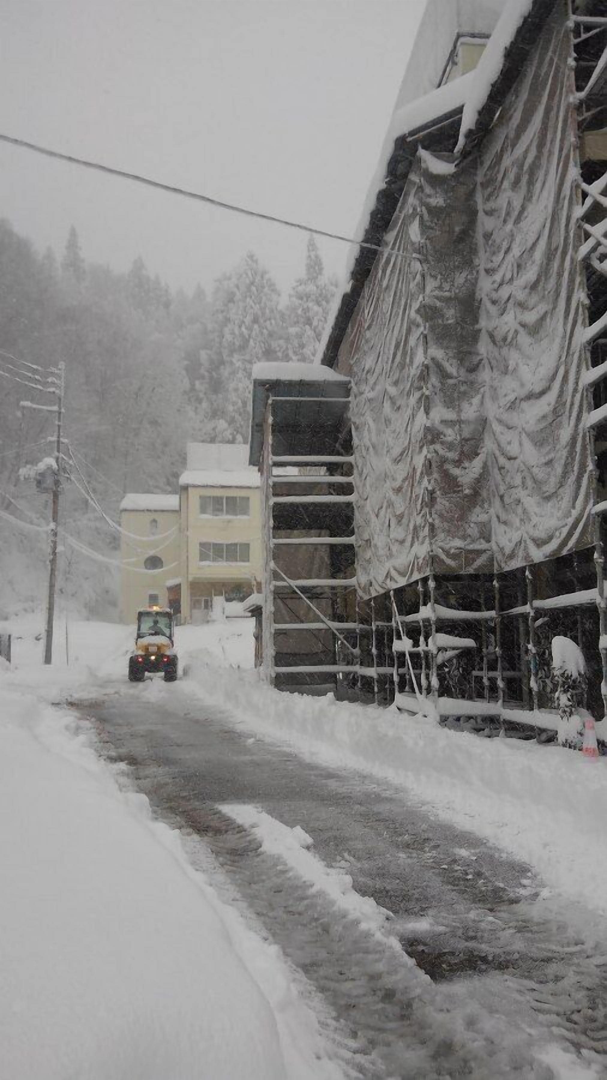 Eine verschneite Straße mit einem Gebäude auf der rechten Seite, einem Fahrzeug in der Ferne und einem teilweise sichtbaren gelben Gebäude im Hintergrund.