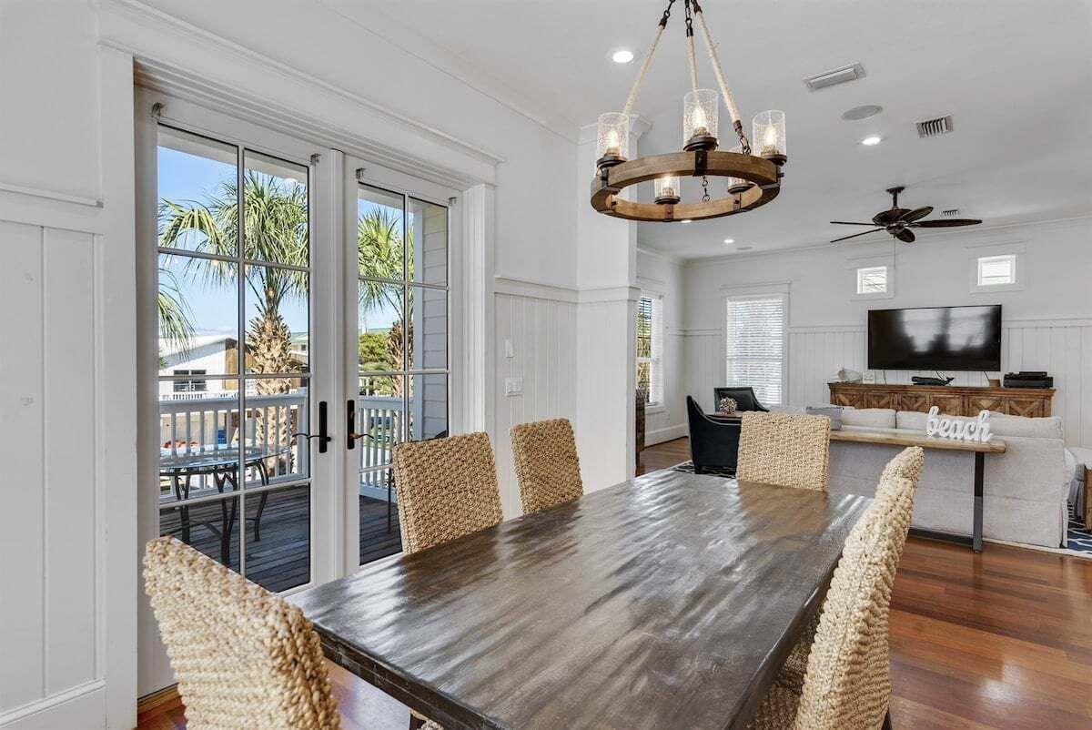 A dining area with a large wooden table and woven chairs, a chandelier, and a view of the outdoors through glass doors.