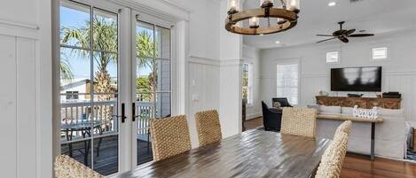 A dining area with a large wooden table and woven chairs, a chandelier, and a view of the outdoors through glass doors.