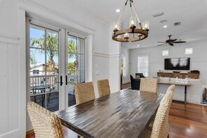 Un espace repas avec une grande table en bois et des chaises en osier, un lustre et une vue sur l’extérieur grâce à des portes vitrées.