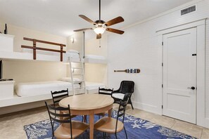 A compact room with bunk beds, a ceiling fan, a small dining table with chairs, and a white door.