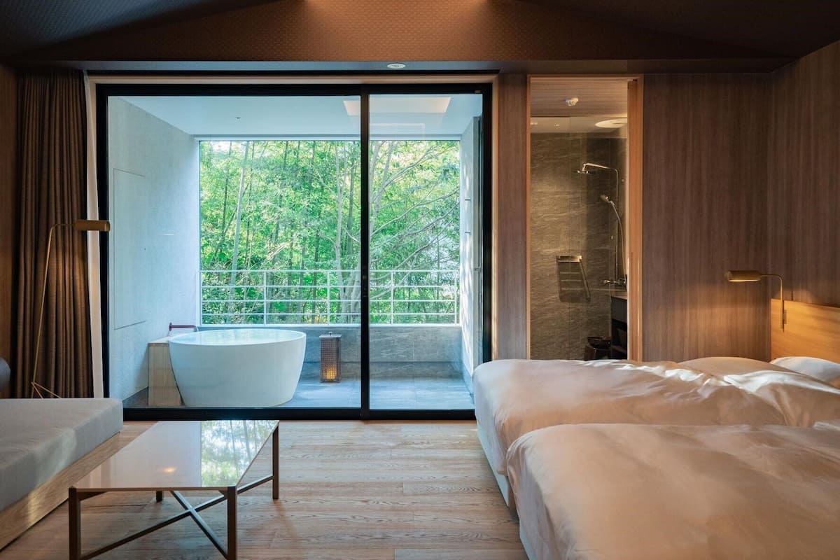 A modern bedroom with a large bed, a sitting area with a table, and a sliding glass door leading to a balcony with a view of trees.