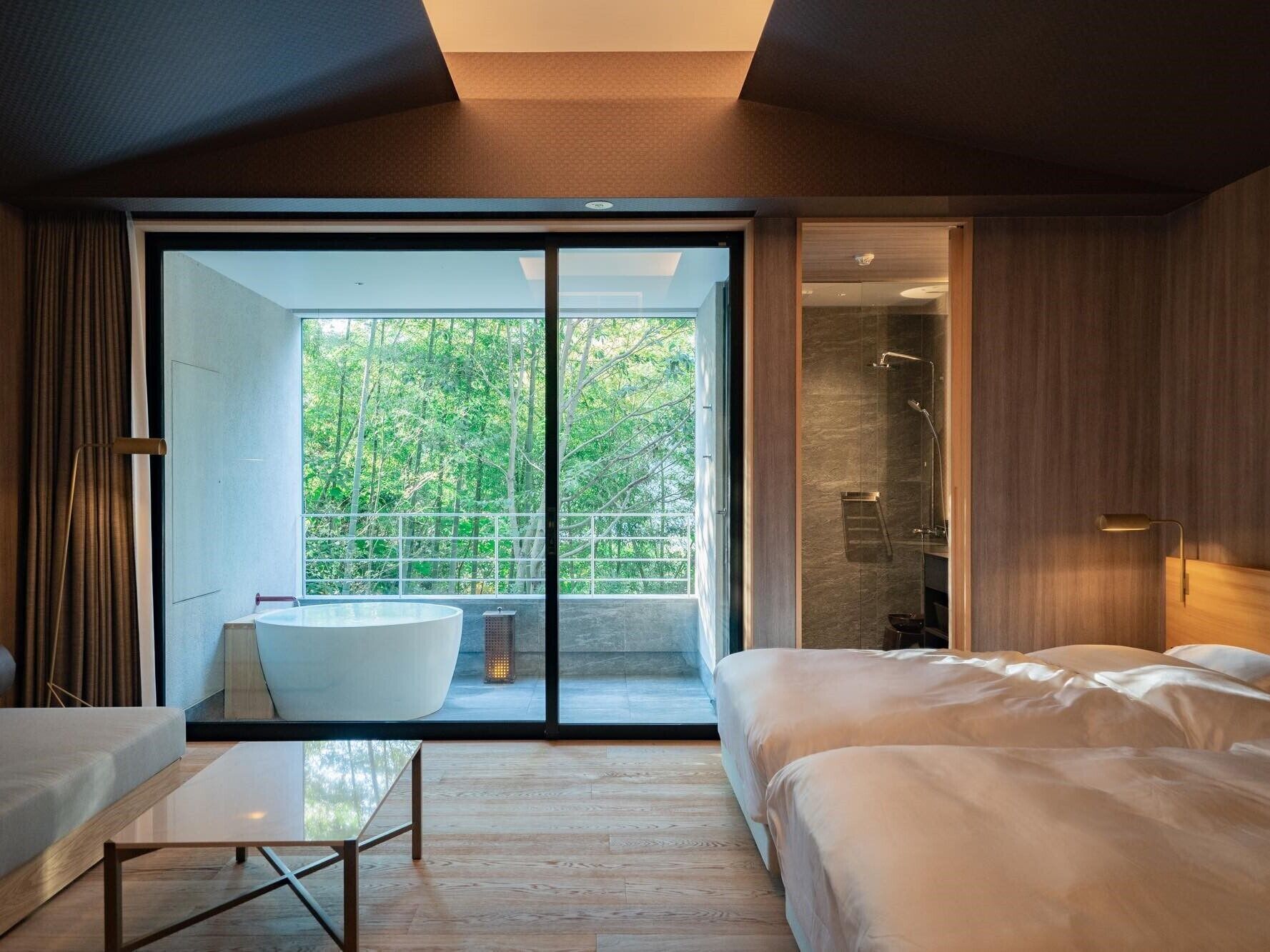 Un dormitorio moderno con una cama grande, una zona de estar con una mesa, una bañera y un ventanal corredero que da a un balcón con vistas a árboles.