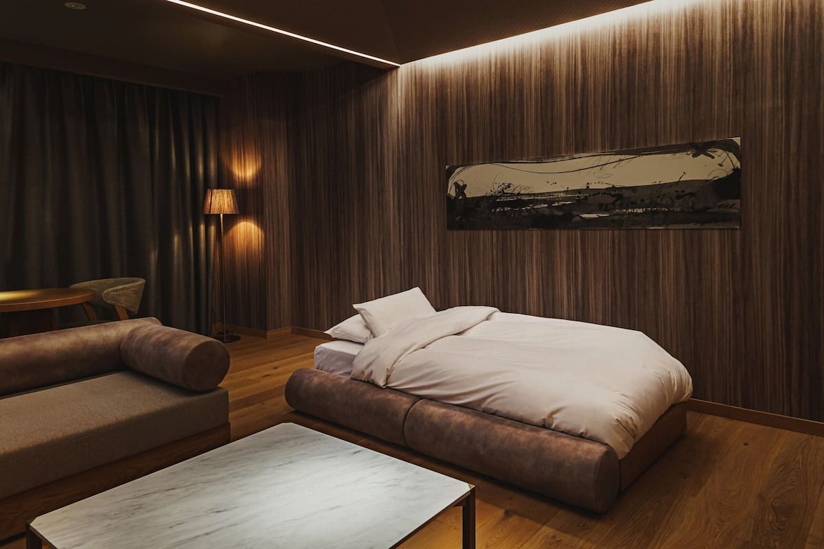 A modern hotel room with a bed, sofa, table, lamp, and artwork on the wall.