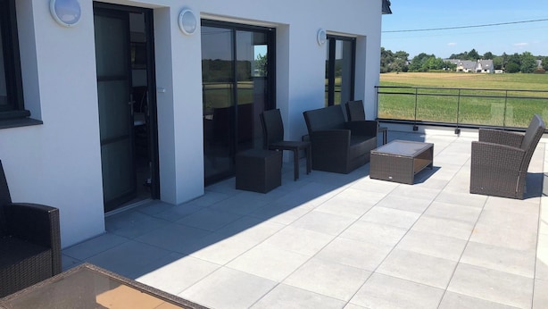 A modern outdoor patio with wicker furniture, glass doors leading to a covered area, and a view of a green field. - Homerez - House in Baden (Baden)